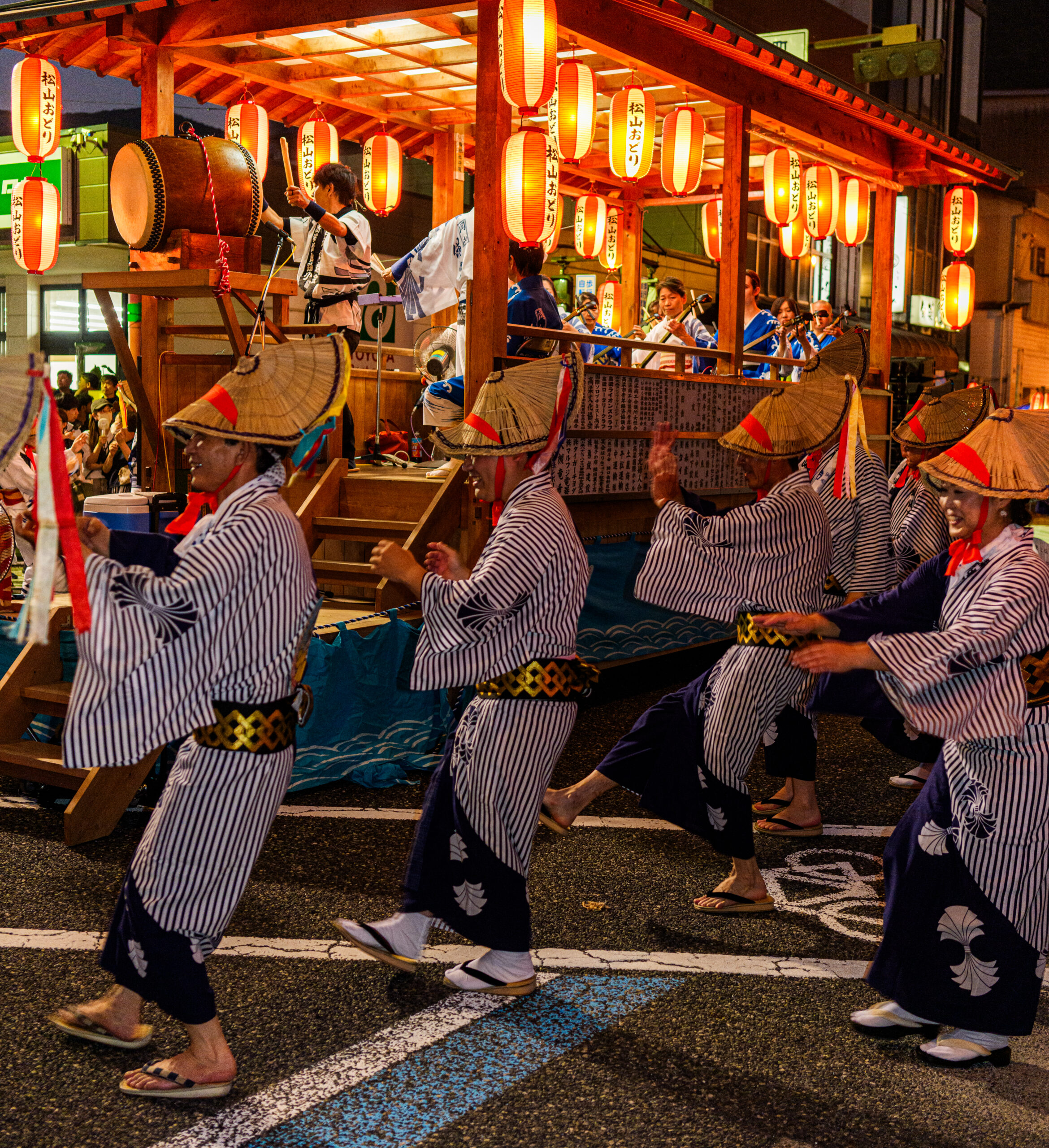 祭りの山車と、その前で踊る踊り手たちの姿。太鼓を叩く人々や、踊りの動きが生き生きと描写されている。